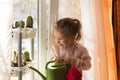 A little girl with a green watering can is playing on the window of the house and watering the flowers. Royalty Free Stock Photo