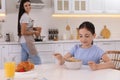 Little girl having breakfast while mother cooking food in kitchen. Single parenting Royalty Free Stock Photo