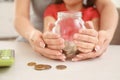 Little girl with her mother sitting at table and holding glass jar with coins, closeup. Money savings concept Royalty Free Stock Photo