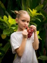 Little girl holding bunch of red grapes, Caucasian girl eating grape. Selected focus. Green tropical leaves background. Organic