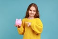 Little girl holding and pointing at piggy bank with finger, suggesting you to put coins inside. Royalty Free Stock Photo