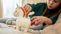 A little girl holding a red tail of her porcelian unicorn moneybox and putting a coin into a slot while sitting on a
