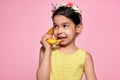 A little girl holds a banana, like a phone, isolated on pink background. Playful kid in a yellow dress pretends to talk on a