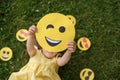 A little girl holds a paper cute winking emoticon lying on the grass of a lawn in the park