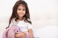 Little girl in home wear sits with teddy bunny on bed at home