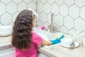 Little girl in household gloves washing up tableware with foamy dishwashing sponge in kitchen sink under water jet.