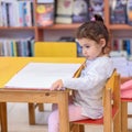 Little Girl Indoors In Front Of Books. Cute Young Toddler Sitting On A Chair Near Table and Reading Book. Royalty Free Stock Photo