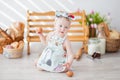 A little girl in a kitchen apron and a bow on her head sits near a bench, next to a basket with buns, bread and donuts Royalty Free Stock Photo