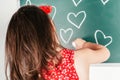 Little girl with long curly hair in red clothes standing back, drawing hearts with chalk on green blackboard. Close up. Royalty Free Stock Photo