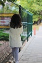 A little girl with long dark hair in a fluffy white coat turned away near the metal fence. Blurred back street background Royalty Free Stock Photo