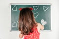 Little girl with long hair in red clothes standing back, drawing hearts with chalk on green blackboard Royalty Free Stock Photo