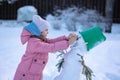 Little girl makes snowman on winter frosty day. Child makes nose out of carrot. Royalty Free Stock Photo