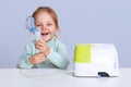 Little girl making inhalation with nebulizer at home, child suffers from asthma, uses inhaler, sits at white desktop, looks Royalty Free Stock Photo