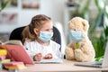 Little girl in mask, with Teddy bear, doing homework, e-learning Royalty Free Stock Photo