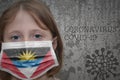 Little girl in medical mask with flag of antigua and barbuda stands near the old vintage wall with text coronavirus, covid, and Royalty Free Stock Photo