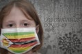 Little girl in medical mask with flag of zimbabwe stands near the old vintage wall with text coronavirus, covid, and virus picture Royalty Free Stock Photo