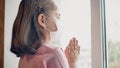 A little girl in medical mask pray for good health near the window