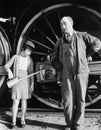 Little girl with an oil can standing next to a locomotive and the engine driver