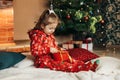 A little girl opens a Christmas present near the Christmas tree. Christmas time, gifts, festive mood. The concept of Royalty Free Stock Photo