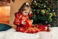 A little girl opens a Christmas present near the Christmas tree. Christmas time, gifts, festive mood. The concept of Royalty Free Stock Photo
