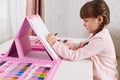 Little girl painting while sitting at desk, making illustration via set of creativity and art, side view of small cute artist with