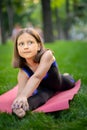 A little girl in the park is doing yoga stretching leaning on her leg. A girl is sitting on a twine