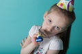 Little girl with party hat on mint background. Birthday girl blowing into party horn blower with hat on.