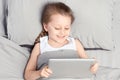 A little girl with a pigtail is lying in bed with a tablet. Solid grey bed linen close-up view from above