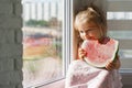 Little girl in a pink dress sits on the window and eats a large piece of watermelon Royalty Free Stock Photo