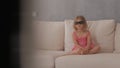 A little girl in a pink dress sitting on the couch attentively and emotionally watching TV in 3D glasses