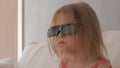 A little girl in a pink dress sitting on the couch attentively and emotionally watching TV in 3D glasses