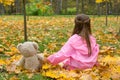 A little girl in a pink raincoat is holding a teddy bear behind her paw in the autumn.