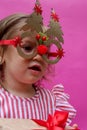 A little girl in a pink striped dress with Christmas tree-shaped glasses. The girl& x27;s very surprised festive face