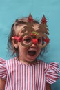 A little girl in a pink striped dress with Christmas tree-shaped glasses. The girl& x27;s very surprised festive face