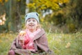 Little girl in a plaid sitting in the autumn park. A child with an apple in his hand is sitting on the grass. A girl wrapped in a Royalty Free Stock Photo