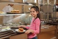 Little girl with plastic tray and burger near serving line in canteen