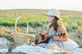 Little girl play ukulele sitting on blanket on field. Portrait of child in panama hat. Outdoor picnic with food basket. Royalty Free Stock Photo