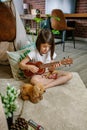 Little girl playing ukulele sitting on carpet next to handmade shelter tent in living room at home Royalty Free Stock Photo