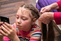 A little girl plays on her phone while her mother does her hair. A woman braids a little girl`s hair. A woman combs a child Royalty Free Stock Photo