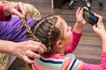 A little girl plays on her phone while her mother does her hair. A woman braids a little girl`s hair. A woman combs a child Royalty Free Stock Photo