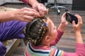 A little girl plays on her phone while her mother does her hair. A woman braids a little girl`s hair. A woman combs a child Royalty Free Stock Photo