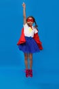 Little girl plays superhero jumping up. Kid on the background of bright blue wall. Girl power concept