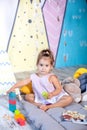 Little girl plays wigwam with colorful toy bricks and builds tower. Happy baby plays in tent in children`s room with educational t Royalty Free Stock Photo