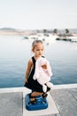 Little girl with a plush hare sits on a bollard on a pier by the sea