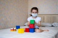 A little girl in a protective mask plays with educational toys, sitting on the bed in self-isolation, look at camera Royalty Free Stock Photo