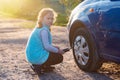 A little girl pumps up a car wheel with a compressor at sunset on a dirt road Royalty Free Stock Photo