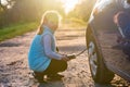 A little girl pumps up a car wheel with a compressor at sunset on a dirt road Royalty Free Stock Photo