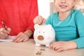 Little girl putting coin into piggy bank and her mother at table. Saving money Royalty Free Stock Photo