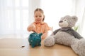 Little girl putting coin into piggy bank for saving with pile of coins on table at home. Royalty Free Stock Photo