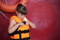 Little girl putting on orange life vest near red wall with safety ring. Space for text Royalty Free Stock Photo